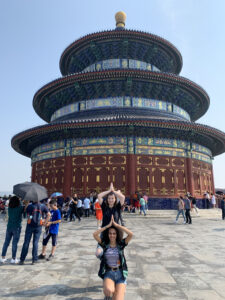 Two people posing in from of the Temple of Heaven in Beijin, China. The building is tall, blue and red with round features. There are tourists in the background