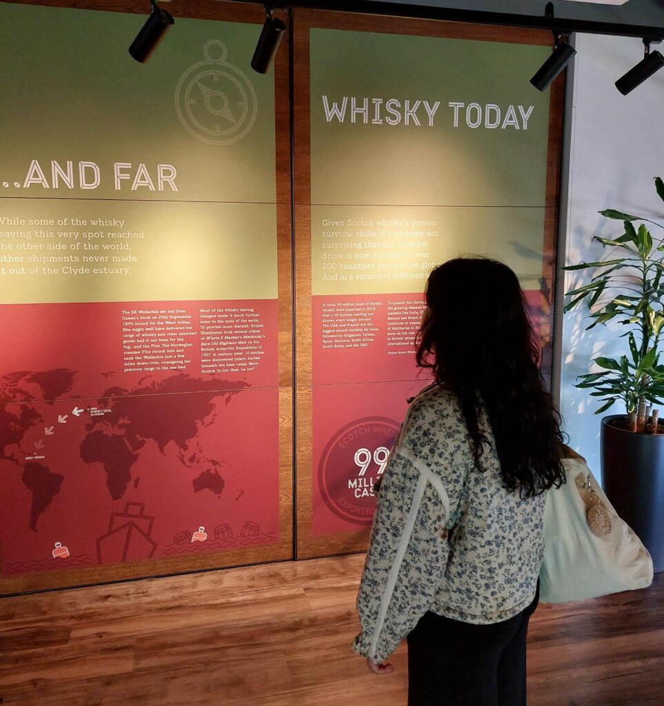 Person with long hair standing with their back turned. They are reading a red and gold poster about whisky.