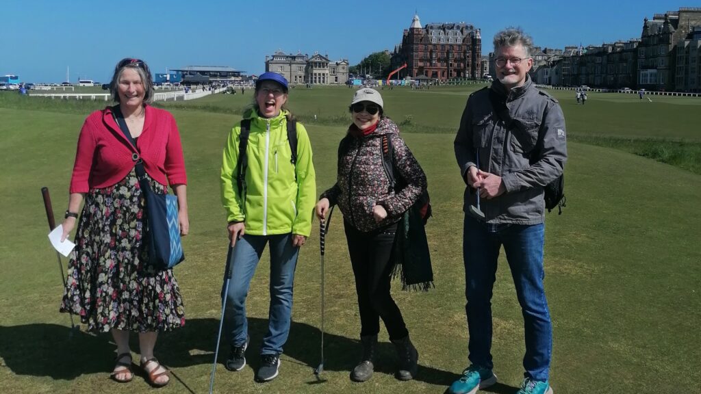 Four staff stand with golf clubs, with the 18th hole and St Andrews landmarks behind them.