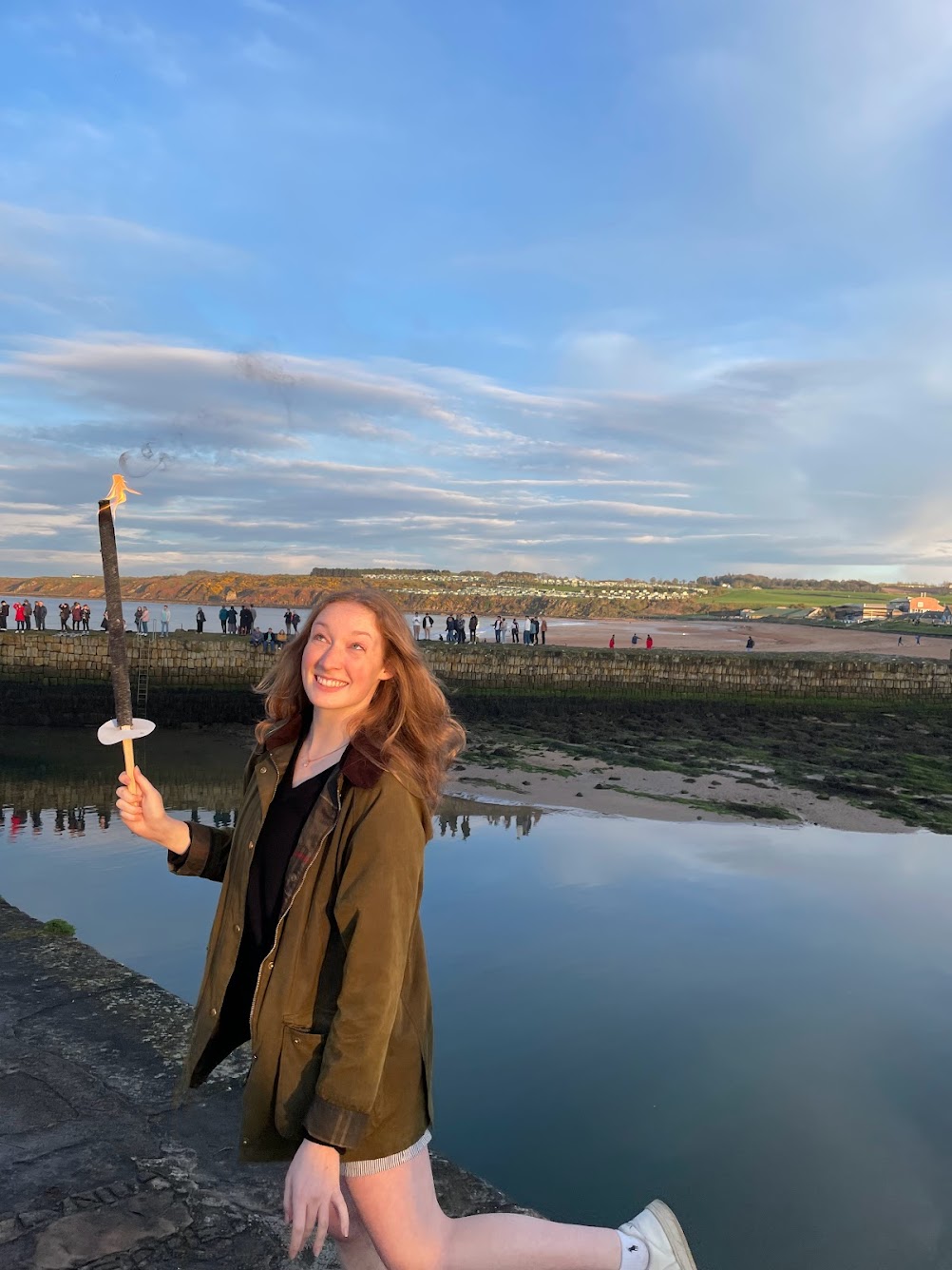 Smiling and looking up into the evening sky, Arden is holding up a lit torch, with the St Andrews pier visible in the background and a number of students walking along to enjoy the views onto the calm sea.
