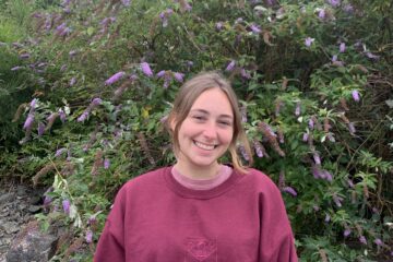 Lauren is standing in front of green and purple bush wearing a maroon St Andrews University jumper.
