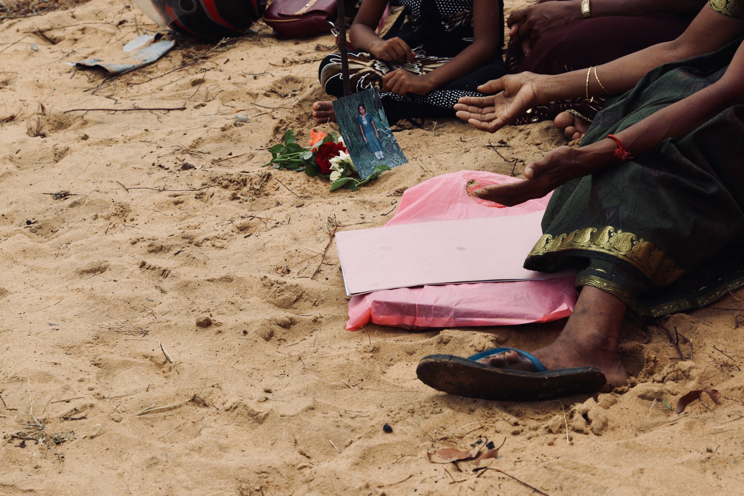 Photograph of a family mourning, with a studio photo of a deceased family member. The people are sitting on sand. There are hands outstrethced towards the photgraph.