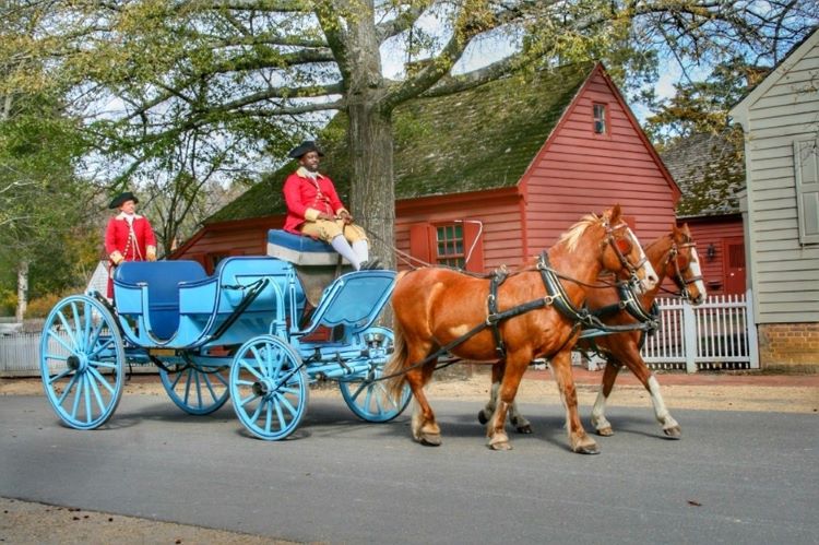 Two men dressed in colonial American hats and red jackets ride a blue horse-drawn carriage.